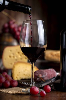 Bir bardak kırmızı şarap ve ahşap masada servis edilen bir şişe peynir, ekmek ve salam. Taze üzüm, bıçak ve tahta. Durgun fotoğraf.