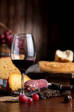 Bir bardak kırmızı şarap ve ahşap masada servis edilen bir şişe peynir, ekmek ve salam. Taze üzüm, bıçak ve tahta. Durgun fotoğraf.