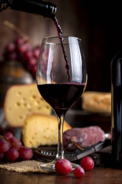 Bir bardak kırmızı şarap ve ahşap masada servis edilen bir şişe peynir, ekmek ve salam. Taze üzüm, bıçak ve tahta. Durgun fotoğraf.
