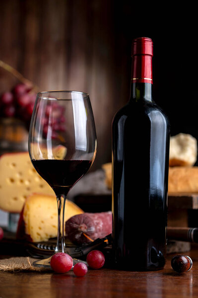 Glass of red wine with a bottle serving on a wooden table with cheese, bread, and salami. Fresh grapes, a knife, and a board. Still life photo.