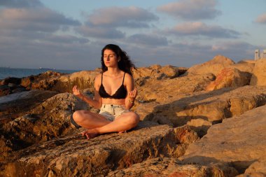 Siyah bikinili uzun saçlı güzel esmer kız gün batımında sahilde kayalıklarda oturuyor..