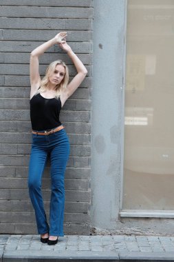 Kot pantolonlu ve çizgili bluzlu güzel sarışın manken büyük bir gülümsemeyle eski bir binanın önünde poz veriyor.