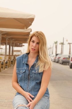 Siyah gömlekli ve kot pantolonlu bir sarışın bir kafede bankta oturuyor.