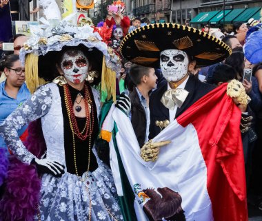 Kız Catrina gibi giyinmiş, adam Charro kafatası gibi giyinmiş, Meksika bayrağını Dia de Muertos Geçit Töreni 'nde tutuyor, Mexico City' de.