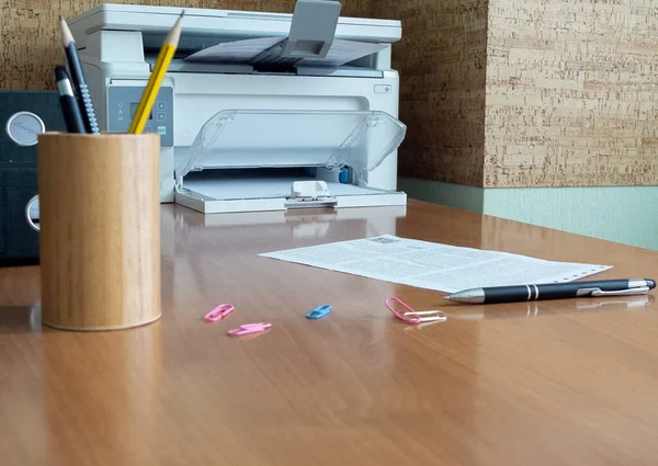 A sheet of paper and a pencil lying on a table against a background of office supplies. A printer is in the background