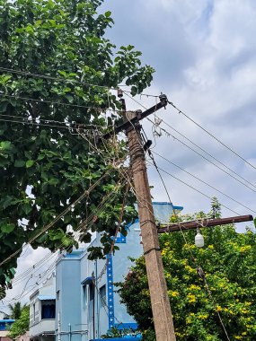 Arkasında yeşil ağaçlar ve bir apartman binası bulunan birbirine dolanmış kablolar olan elektrik direği. Kablolar, yakınlardaki ağaç dallarıyla iç içe geçmiş gibi görünüyor. Bulutlu bir gökyüzünün altında karmakarışık bir manzara yaratıyor..