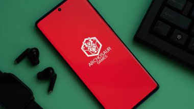 Dhaka, Bangladesh 01 June 2025:Red screen smartphone with Archosaur Games logo among tech gadgets.