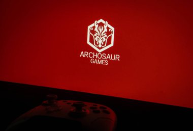 Dhaka, Bangladesh 01 June 2025:Archosaur Games logo on a red screen with a gaming controller in view.