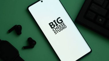 Dhaka, Bangladesh 02 June 2025:Smartphone with BIG CHEESE STUDIO logo, earbuds, and smartwatch on green surface.