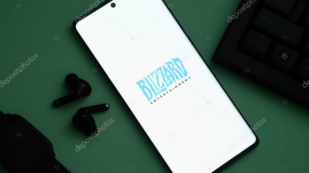 Dhaka, Bangladesh 02 June 2025:Smartphone showing BLIZZARD ENTERTAINMENT logo with earbuds and smartwatch on green.