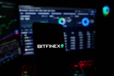 Dhaka, Bangladeş 18 Eylül 2025: Şifreli telefon üzerindeki Bitfinex logosu ve arkasında para birimleri ve finansal ticaret grafikleri