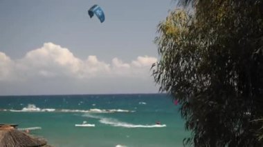 Turkuaz okyanus dalgaları üzerinde heyecanlı uçurtmalar. Parlak gökyüzünün altında saman şemsiyeli sakin bir sahilde..