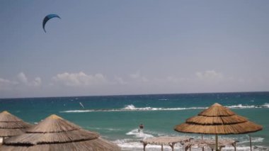 Turkuaz okyanus dalgaları üzerinde heyecanlı uçurtmalar. Parlak gökyüzünün altında saman şemsiyeli sakin bir sahilde..