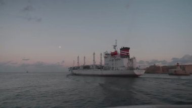 Alacakaranlıkta sakin okyanus sularında hareket eden büyük bir kargo gemisi canlı deniz hareketlerini yansıtıyor.