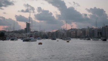 Limandaki huzurlu ve sakin bir akşam sahnesinde yelkenli tekneleri ve bulutlu gökyüzünün altında şehir silueti yer alıyor.