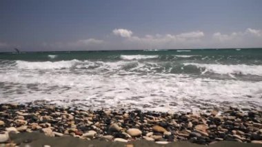 Dalgalar bir çakıl taşlı plaja çarpar berrak, güneşli bir gökyüzünün altında uzak ufuklara bakan yelkenli tekneleriyle.