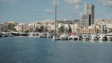 Resimli bir marina, sakin sularda yüzen demirli yatları şehrin ufuk çizgisine karşı sergiliyor.