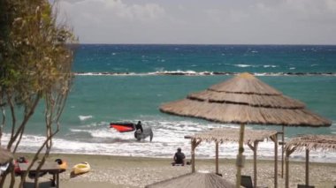 Turkuaz okyanus dalgaları üzerinde heyecanlı uçurtmalar. Parlak gökyüzünün altında saman şemsiyeli sakin bir sahilde..