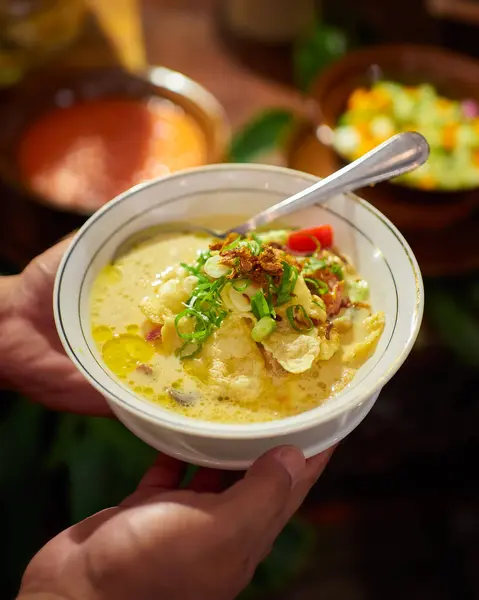 Bir kişi geleneksel bir Endonezya et çorbası olan Soto Betawi 'yi hindistancevizi sütü çorbasının üzerine kızarmış soğan, soğan ve kaşık ile servis eder.