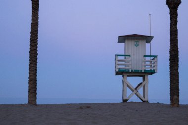 Alacakaranlıkta iki palmiye ağacı tarafından çerçevelenmiş kumlu bir plajın üzerinde beyaz ve deniz mavisi cankurtaran kulesi yükseliyor. Seyahat, yaz veya plaj temalı görüntüler