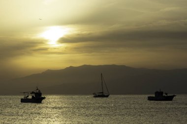 Sakin deniz manzarası, altın bir günbatımına karşı silueti olan üç tekne, sakin sularda, dağlık bir zemin üzerinde usulca süzülüyor. Sakin atmosfer ve yumuşak ılık tonlar, doğayla ilgili kavramlar için ideal bir huzur ve dinginlik hissi uyandırıyor.