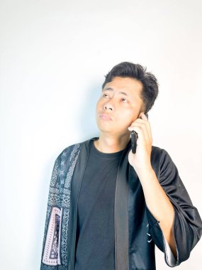 Düşünceli bir yüz ifadesi olan genç bir adam cep telefonuyla konuşuyor. Geleneksel desenleri olan siyah bir kıyafet giyiyor, ciddi ve odaklanmış görünüyor. Temiz beyaz arka plan, minimalist ve profesyonel bir izlenim veriyor. Komün kavramları için ideal