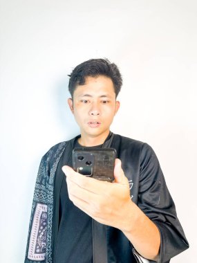 Ciddi bir ifadesi olan genç bir adam telefonuna odaklanmış durumda. Japon harf aksanlı siyah bir ceket giyiyor, minimalist beyaz bir arka plana karşı duruyor. Teknoloji, iletişim ya da dijital yaşam tarzı kavramları için uygundur.