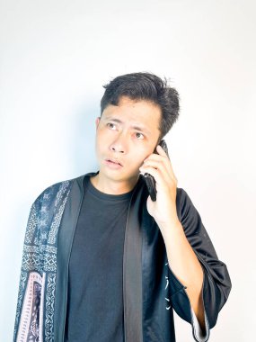 Düşünceli bir yüz ifadesi olan genç bir adam cep telefonuyla konuşuyor. Geleneksel desenleri olan siyah bir kıyafet giyiyor, ciddi ve odaklanmış görünüyor. Temiz beyaz arka plan, minimalist ve profesyonel bir izlenim veriyor. Komün kavramları için ideal