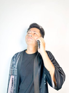 Düşünceli bir yüz ifadesi olan genç bir adam cep telefonuyla konuşuyor. Geleneksel desenleri olan siyah bir kıyafet giyiyor, ciddi ve odaklanmış görünüyor. Temiz beyaz arka plan, minimalist ve profesyonel bir izlenim veriyor. Komün kavramları için ideal