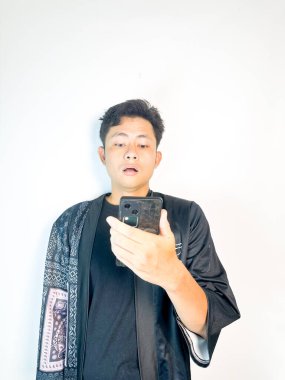 Ciddi bir ifadesi olan genç bir adam telefonuna odaklanmış durumda. Japon harf aksanlı siyah bir ceket giyiyor, minimalist beyaz bir arka plana karşı duruyor. Teknoloji, iletişim ya da dijital yaşam tarzı kavramları için uygundur.