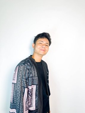 Geleneksel desenli siyah cüppeli genç bir adam başını hafifçe eğerken neşeli bir sırıtış yapar. Yüzündeki ifade eğlence, yaramazlık ya da hafif bir tepki veriyor. Bu da onu mizah, merak ya da iğneleme gibi kavramlar için ideal kılıyor..