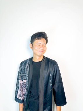 Geleneksel desenli siyah cüppeli genç bir adam başını hafifçe eğerken neşeli bir sırıtış yapar. Yüzündeki ifade eğlence, yaramazlık ya da hafif bir tepki veriyor. Bu da onu mizah, merak ya da iğneleme gibi kavramlar için ideal kılıyor..