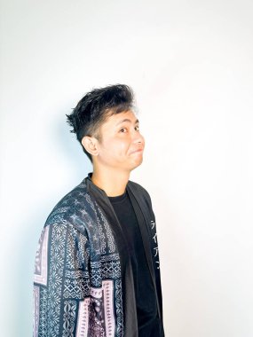 Geleneksel desenli siyah cüppeli genç bir adam başını hafifçe eğerken neşeli bir sırıtış yapar. Yüzündeki ifade eğlence, yaramazlık ya da hafif bir tepki veriyor. Bu da onu mizah, merak ya da iğneleme gibi kavramlar için ideal kılıyor..