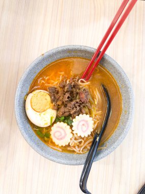 Haşlanmış yumurta, balık köftesi, sebze ve yemek çubuklarıyla sıcak ve baharatlı erişte kasesinin üst görüntüsü. Tahta bir masada servis edilmiş. Japon mutfağı, konforlu yemekler, Asya yemekleri ve erişte çorbası gibi konular için mükemmel..