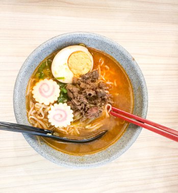 Haşlanmış yumurta, balık köftesi, sebze ve yemek çubuklarıyla sıcak ve baharatlı erişte kasesinin üst görüntüsü. Tahta bir masada servis edilmiş. Japon mutfağı, konforlu yemekler, Asya yemekleri ve erişte çorbası gibi konular için mükemmel..