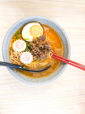 Haşlanmış yumurta, balık köftesi, sebze ve yemek çubuklarıyla sıcak ve baharatlı erişte kasesinin üst görüntüsü. Tahta bir masada servis edilmiş. Japon mutfağı, konforlu yemekler, Asya yemekleri ve erişte çorbası gibi konular için mükemmel..