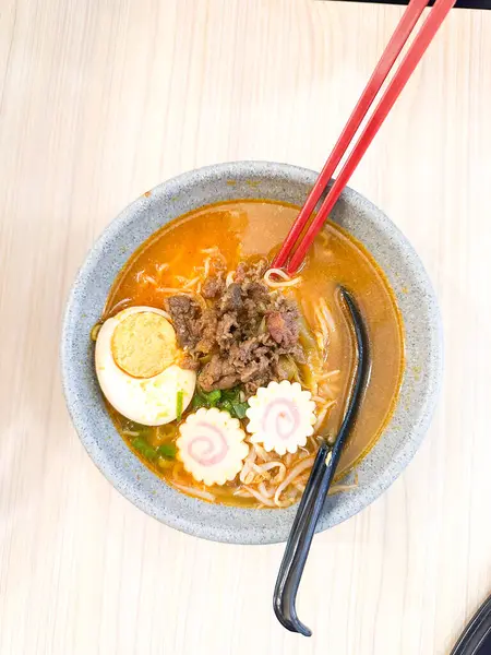 Haşlanmış yumurta, balık köftesi, sebze ve yemek çubuklarıyla sıcak ve baharatlı erişte kasesinin üst görüntüsü. Tahta bir masada servis edilmiş. Japon mutfağı, konforlu yemekler, Asya yemekleri ve erişte çorbası gibi konular için mükemmel..