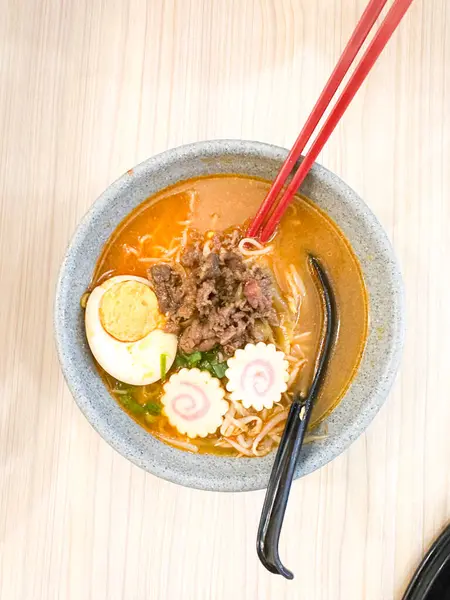 Haşlanmış yumurta, balık köftesi, sebze ve yemek çubuklarıyla sıcak ve baharatlı erişte kasesinin üst görüntüsü. Tahta bir masada servis edilmiş. Japon mutfağı, konforlu yemekler, Asya yemekleri ve erişte çorbası gibi konular için mükemmel..