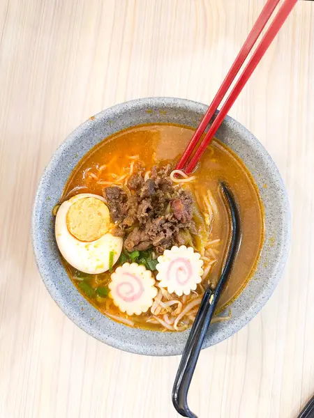 Haşlanmış yumurta, balık köftesi, sebze ve yemek çubuklarıyla sıcak ve baharatlı erişte kasesinin üst görüntüsü. Tahta bir masada servis edilmiş. Japon mutfağı, konforlu yemekler, Asya yemekleri ve erişte çorbası gibi konular için mükemmel..