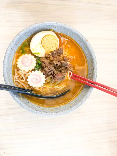 Haşlanmış yumurta, balık köftesi, sebze ve yemek çubuklarıyla sıcak ve baharatlı erişte kasesinin üst görüntüsü. Tahta bir masada servis edilmiş. Japon mutfağı, konforlu yemekler, Asya yemekleri ve erişte çorbası gibi konular için mükemmel..