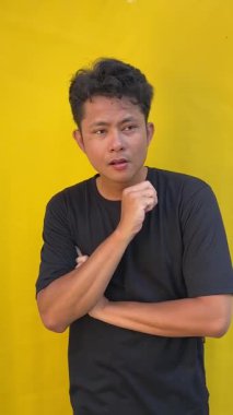 Sarı tişört ve siyah gömlek giyen yakışıklı genç bir adam. Ciddi görünüyor ve el kol hareketi yapıyor. Göğsünde bir şey var.