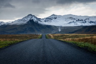 İzlanda kırsalında bulutlu bir günde karlı dağlara doğru giden yol.