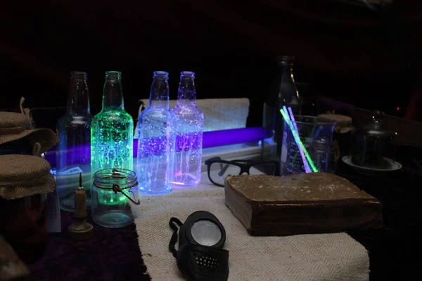 In neon light, on the table there is an alchemists set: jars, glasses, bottles, an old book and other little things.