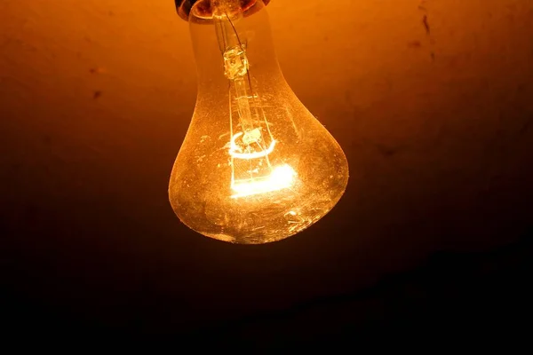 An old, turned on incandescent light bulb hanging from the ceiling.