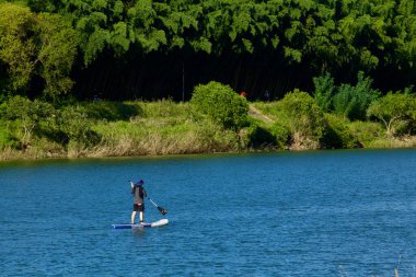 Taehwa Nehri boyunca sakin bir manzara gözler önüne seriliyor, arka planda bambu ormanının huzurlu güzelliği tarafından çerçevelenmiş, suyun üzerinde yavaşça süzülen bir kürekçi..