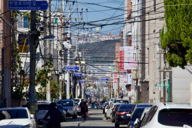 Bu uzun mercek görüntüsü, Ulsan 'da park halindeki arabalarla çevrili ve elektrik hatlarıyla çaprazlanmış tek şeritli bir yan şeritin günlük basitliğini yakalıyor..