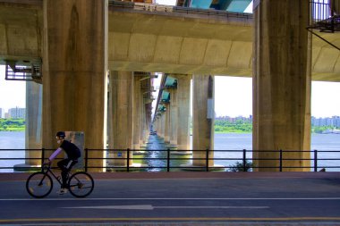Bir bisikletçi, Gangbyeon Otoyolu 'nun altındaki bisiklet yolunda nehrin manzarası ve üzerinden geçen bir metro köprüsü ile şehir ulaşımını ve doğal güzelliği harmanlayan sakin bir yolculuğun keyfini çıkarıyor..