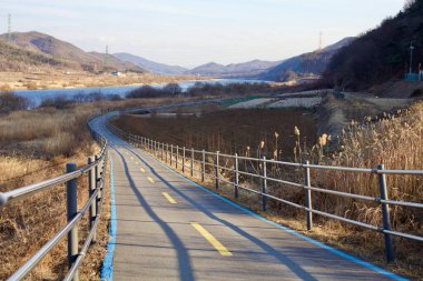 Yeoju Şehri, Güney Kore - 20 Şubat 2020: Uzakta dağları olan sakin Güney Han Nehri 'ne doğru yavaşça eğimli bir bisiklet yolu iner..