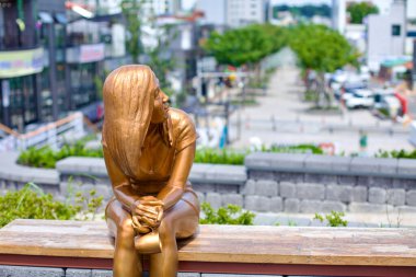 Gangneung Şehri, Güney Kore - 29 Temmuz 2019: Gangneung şehir merkezindeki Wolhwa Caddesi 'ne bakan bir park bankında oturan genç bir kadının altın heykeli elinde bir kupa tutarken sanatsal bir dokunuş ve hareketli bir tasarım içeriyor.