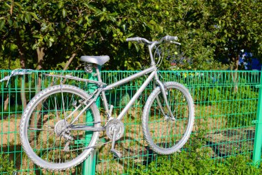 Yangyang County, South Korea - July 30th, 2019: An old, non-functioning bicycle, transformed into a piece of art by being painted entirely gray, hangs off a fence leading to fruit trees near Dongho Beach, symbolizing a creative repurpose. clipart